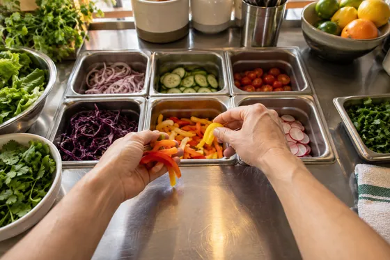 First-person illustration: hands arrange vegetables at a commercial kitchen prep station.